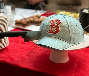 A light blue cake in the shape of a baseball cap sits on a cake stand atop a red table. There is a large 'B' on the front of the cap, and a knife embedded in the cake. On the table are a stack of small plates and some small hamburgers.