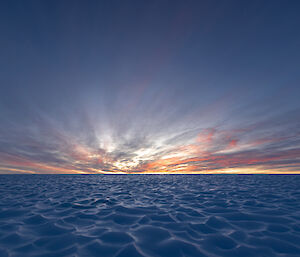 A brightly coloured sunset is in the distance over an vast field of ice