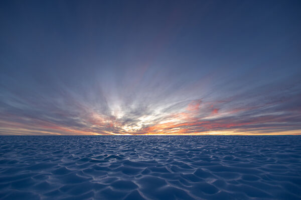 A brightly coloured sunset is in the distance over an vast field of ice