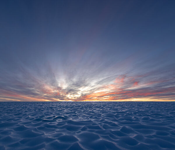 A brightly coloured sunset is in the distance over an vast field of ice