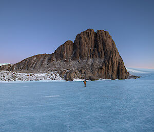 A man is standing on a frozen lake with a rocky peak in the background