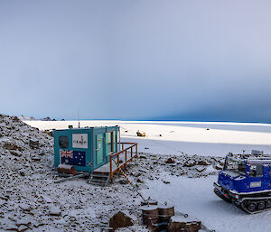 A Hägglunds vehicle is parked on the snow outside a small green hut with the Australian flag painted on the outside. A rocky peak can be seen to the left of frame