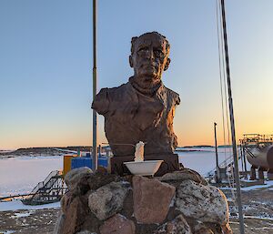 A bowl with noodles frozen solid as if being eaten with the spoon frozen into them sits in front of a bust of Sir Douglas Mawson