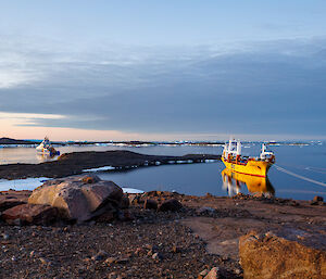 A yellow ship is tied up in a natural harbour while a blue and white ship sits in the water outside the harbour. There are island and icebergs in the distance.