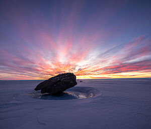 A rock is balanced precariously on the ice with the plateau stretching in all directions. A multi-coloured sunset lights the sky