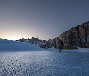 Two men are walking across a frozen lake. There is snow and ice built up on the left of frame and a rocky mountain in the far distance and to the right of frame.