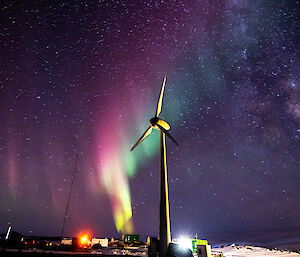 A night photograph of a wind turbine in the foreground with a brightly coloured Aurora stretching across the sky and the milky way visible in the top right of frame