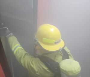 Person wearing a yellow firefighter's uniform and hardhat opening the door to a smoke-filled room.