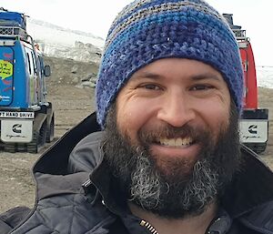 A smiling man wearing a home-made crocheted blue and grey beanie.