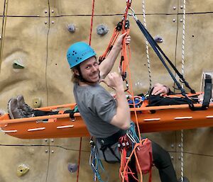 A smiling man wearing a blue hard-hat suspended up a climbing wall while practising the rescue of a patient in a stretcher.