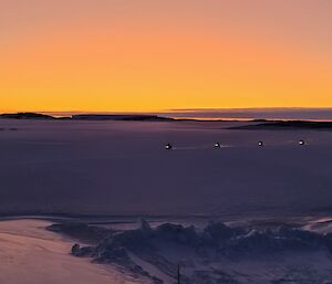 Four quad-bikes are moving across an ice covered harbour with the orange glow of sunset in the distance