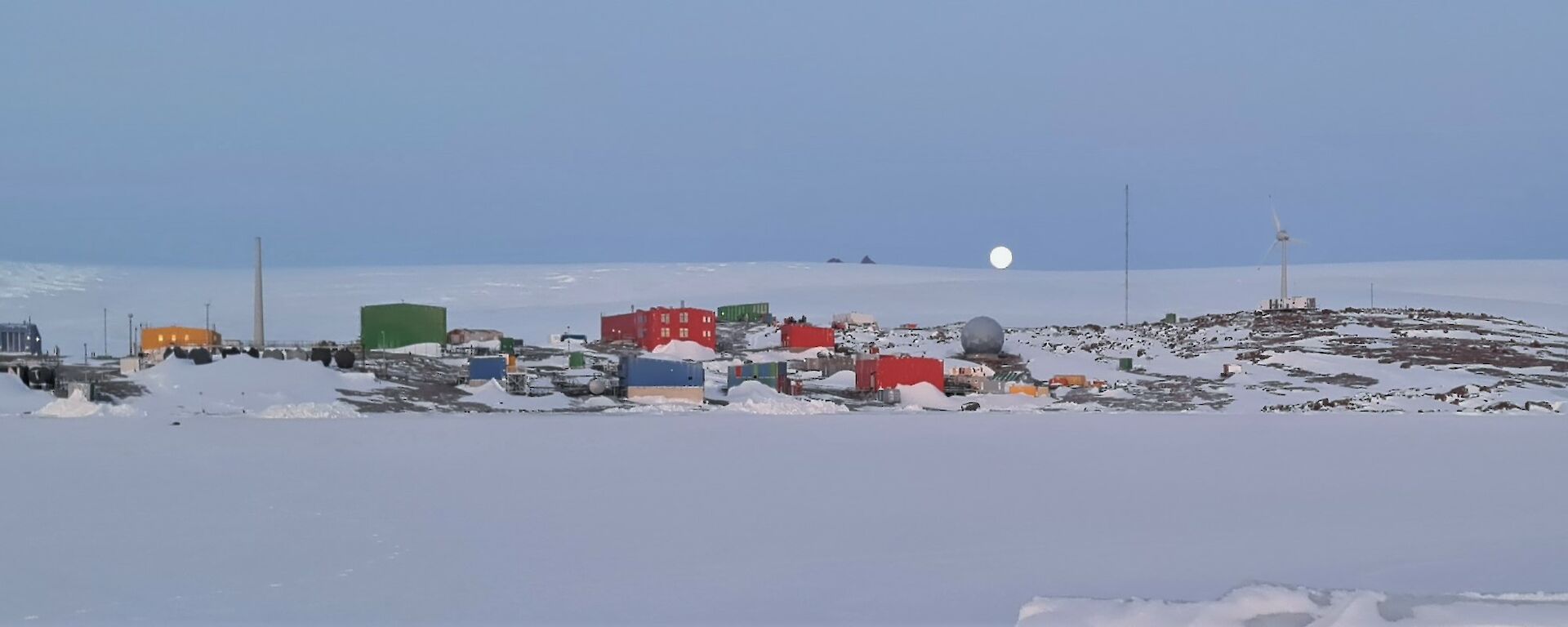Multi coloured buildings lay on a snow covered rocky landscape with a rising ice plateau behind it, sea ice to its front and the moon just on the far horizon