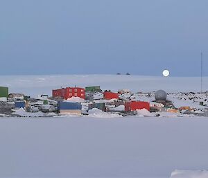 Multi coloured buildings lay on a snow covered rocky landscape with a rising ice plateau behind it, sea ice to its front and the moon just on the far horizon