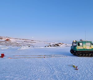 Ice anchors and tow lines are laid out to practice recovery of a green twin cab Hägglunds vehicle on the sea-ice. A wind turbine on a snow covered rocky hill is to the left of frame in the distance
