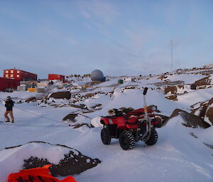 A man is pulling a quad-bike over snow covered rocks with a rope. Brightly coloured buildings, a radome, and a wind turbine are in the background.