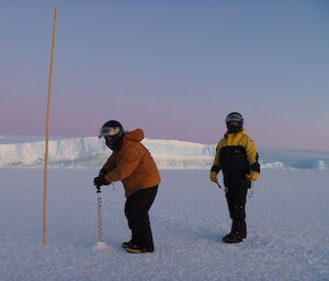 A man is using a drill to drill down into the ice while another man is watching on. Ice cliffs are in the distance
