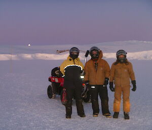 Three men stand posing for the photo in front of a quad-bike on sea-ice. There are ice cliffs in the distance.
