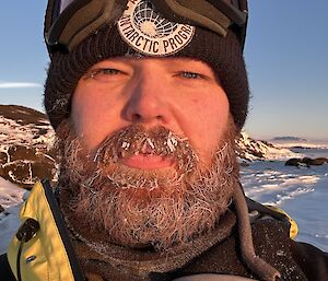 A man looks into the camera. He has ice in his moustache and beard and is wearing a jacket, beanie and goggles on his head,