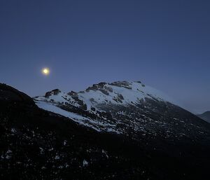 The moon is rising in the distance beyond a snow covered mountain peak