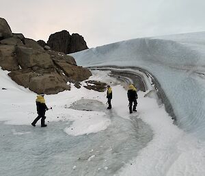 Three men are next to a wind scour formed of ice and snow with a rocky landscape rising to the left of frame