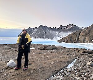 A man is standing on a rocky landscape with a frozen lake in the background and rocky, snow covered mountains in the distance