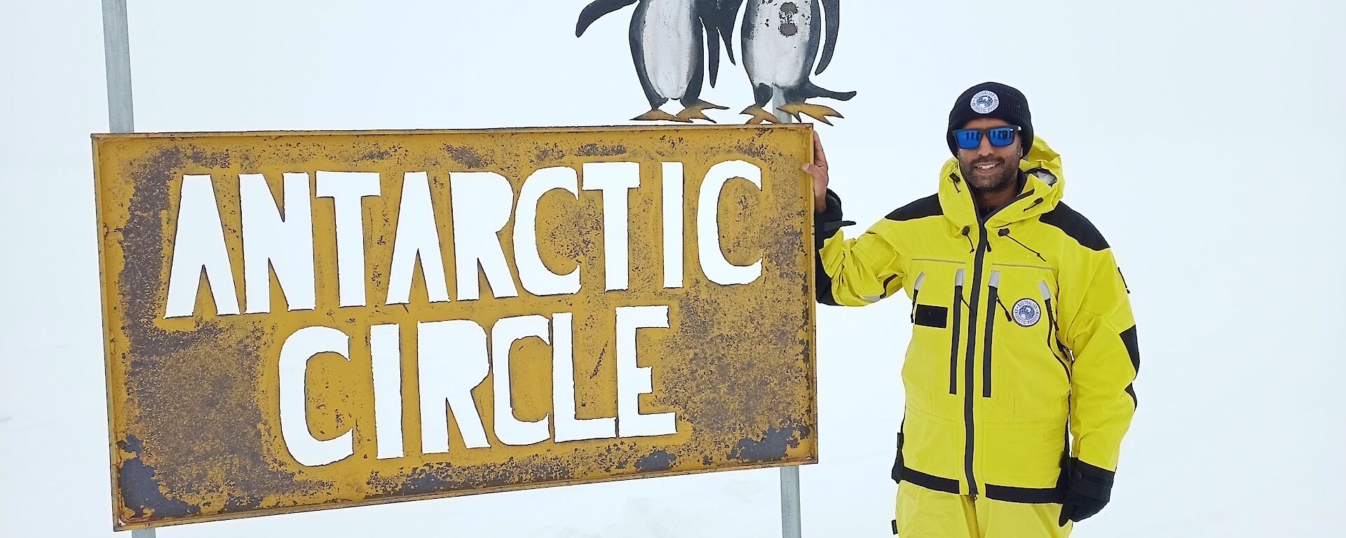 A man in yellow survival clothing stands to the right of a metal sign, with the background completely white with snow. The sign says Antarctic Circle, and has two small metal penguins on the top of it.