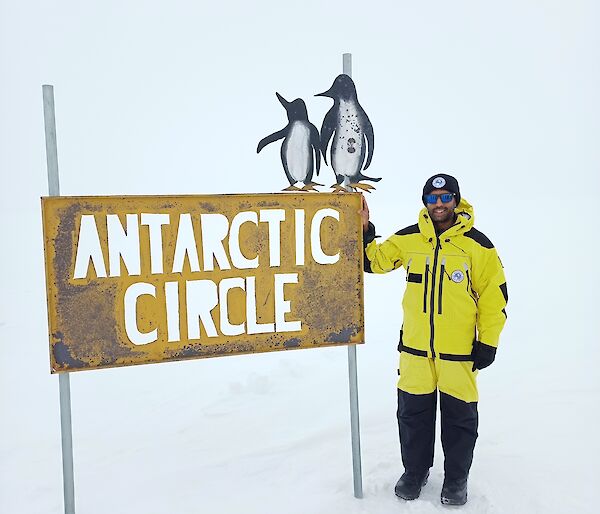 A man in yellow survival clothing stands to the right of a metal sign, with the background completely white with snow. The sign says Antarctic Circle, and has two small metal penguins on the top of it.