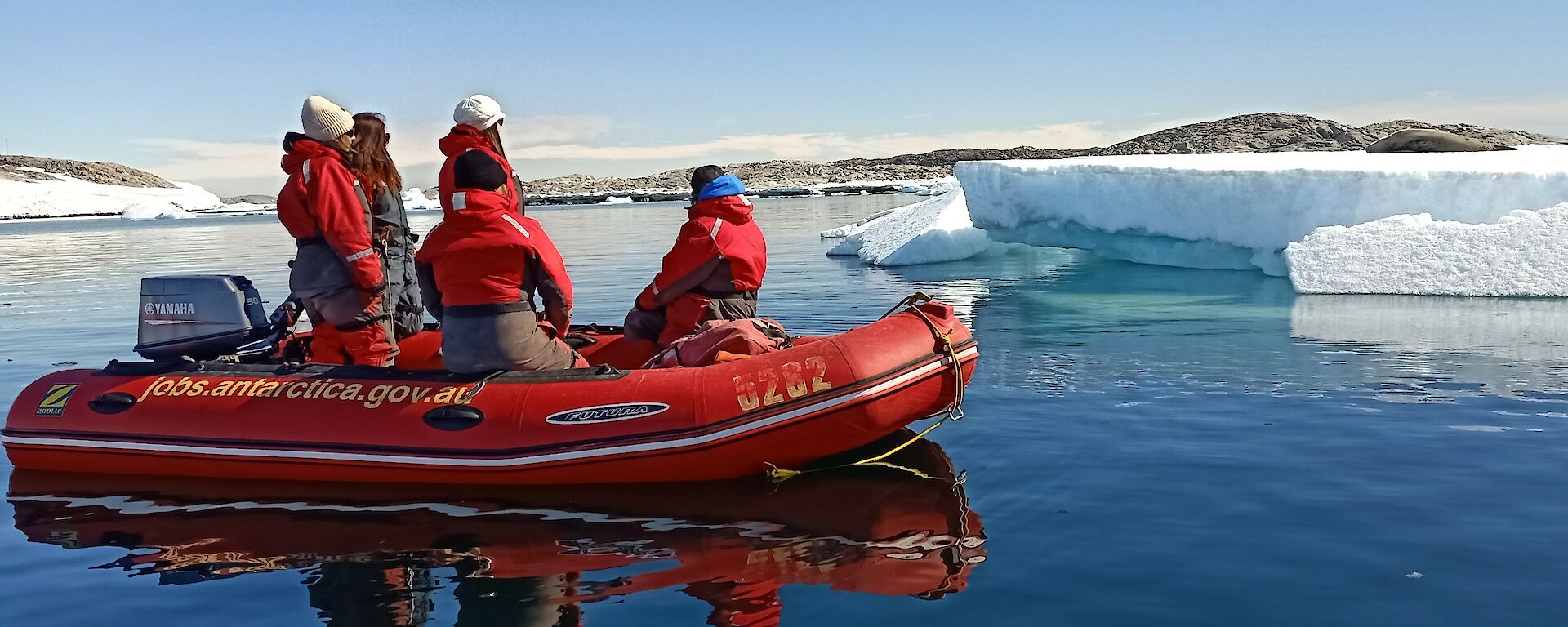A red inflatable boat with five people in it is stationary on some calm flat water. There are several large pieces of ice in the background, and a leopard seal is relaxing on one to the right of frame.