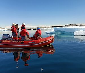 A red inflatable boat with five people in it is stationary on some calm flat water. There are several large pieces of ice in the background, and a leopard seal is relaxing on one to the right of frame.