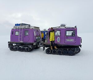 The background of this photo is completely white with snow and a cloudy sky. It is difficult to discern the horizon. There is a purple tracked vehicle in the centre of the shot. The vehicle has two very boxy sections, and a man in yellow survival clothing stands on the tracks of the front section.