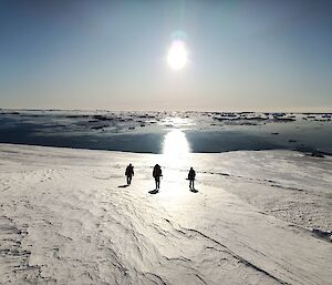 Three people walk across a snow and ice covered ground, away from the camera and into a glaring sun. There is a band of water beyond the snow covered ground and the sky is clear and blue. The sun is low in the sky and reflecting across the water.