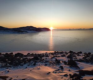 The sun is just sinking below the horizon which stretches across the top third of the photo. The foreground is snow and rock covered ground, with the middle third of the photo being flat calm water. The sun is reflecting across the water towards the camera.