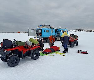 A training scene for the search and rescue team is pictured here. There is a four wheel motorbike to the left of frame and behind that on the ground is a patient on a stretcher. Two men stand either side of the stretcher. In the background is a light blue tracked vehicle. The vehicle has two box shaped sections. The sky is overcast and the ground is covered in snow.