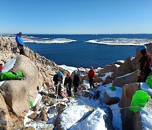 Another training exercise is captured in this photo. The view is looking down a shallow rocky valley, towards some calm blue water and two large snow covered islands. There are men on both sides of the dip observing several other men connecting up ropes to recover a patient from the base of the hill. Everyone is wearing helmets and climbing harnesses. The terrain is made up of various size boulders with a light covering of snow. The sky is clear and blue.