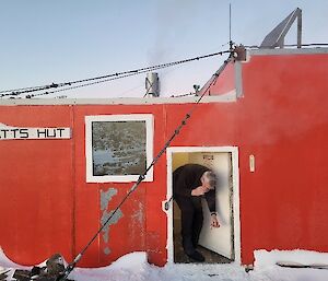 A man standing in the doorway of a red field hut is bent over, looking for something.