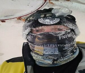 A man wearing a beanie and buff with ice crystals visible on the edges of both as well as on his eyelashes.