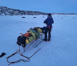 A man standing on the sea-ice hauling a sled fully laden with bags and equipment.