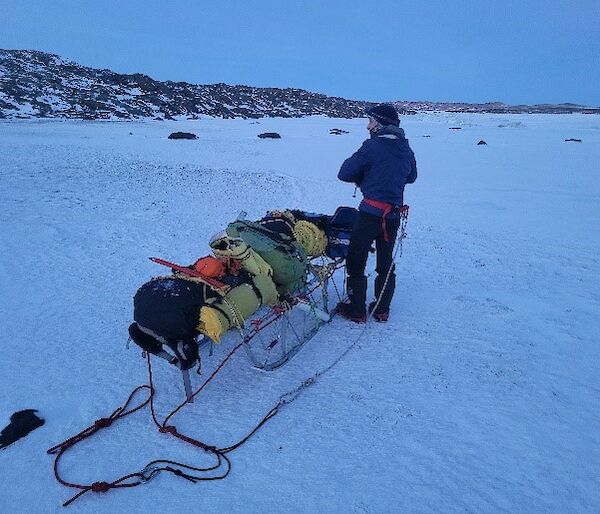 A man standing on the sea-ice hauling a sled fully laden with bags and equipment.