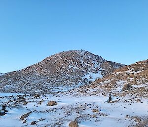 A snow covered hill in the background with blue skies above. The foreground is mostly covered in snow with a few rocks poking through.