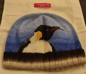A beanie lays on a cloth bag. The beanie is decorated with an image of an Emperor Penguin