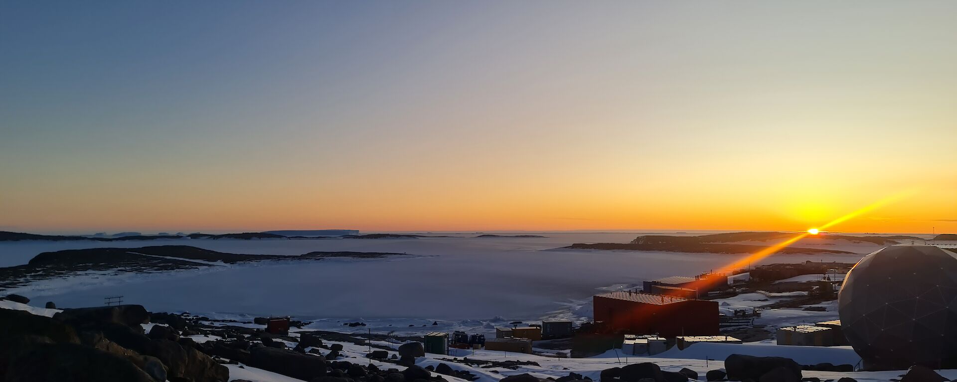 The sun is just on the horizon with an orange glow spreading over islands in the distance in an ice covered sea. Station buildings are to the right of frame and a satellite dome is in the front right of frame.