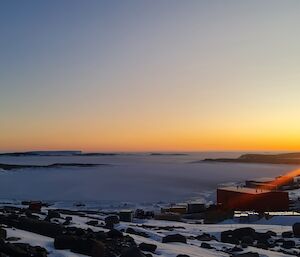 The sun is just on the horizon with an orange glow spreading over islands in the distance in an ice covered sea. Station buildings are to the right of frame and a satellite dome is in the front right of frame.