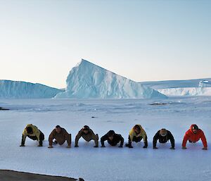 Six men and one woman are doing push-ups on ice with two large ice-bergs in the background and ice-cliffs in the distance.