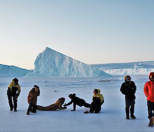 Two men are in a push-up position on ice, with a woman and two other men looking on and two men walking away. There are ice-bergs in the background.