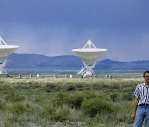 A man standing in a grassy field with three satellite dishes behind.