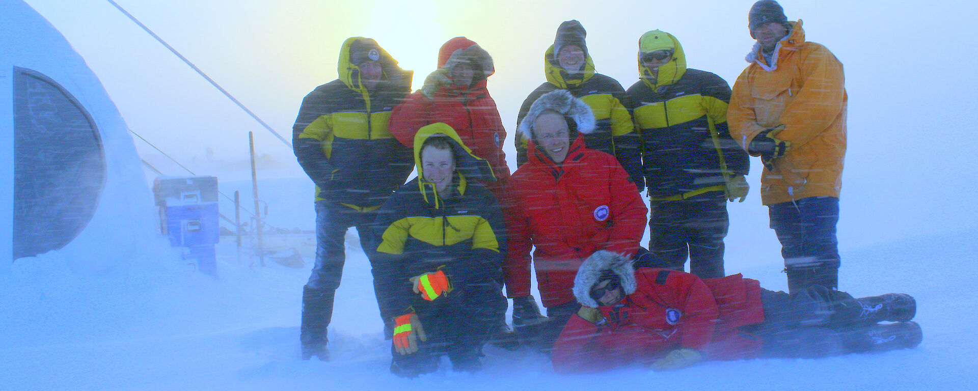 Eight people wearing down jackets posing for the camera during an Antarctic blizzard.
