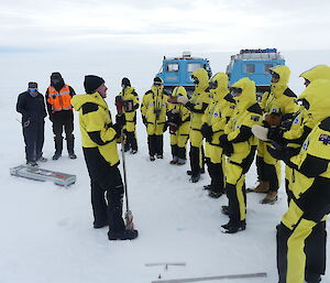A man speaking in front of a microphone in Antarctica, to a group wearing Antarctic yellow and black jackets.