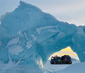 A blue Hagglunds is visible through the arch of a jagged blue and white iceberg set in the sea-ice.