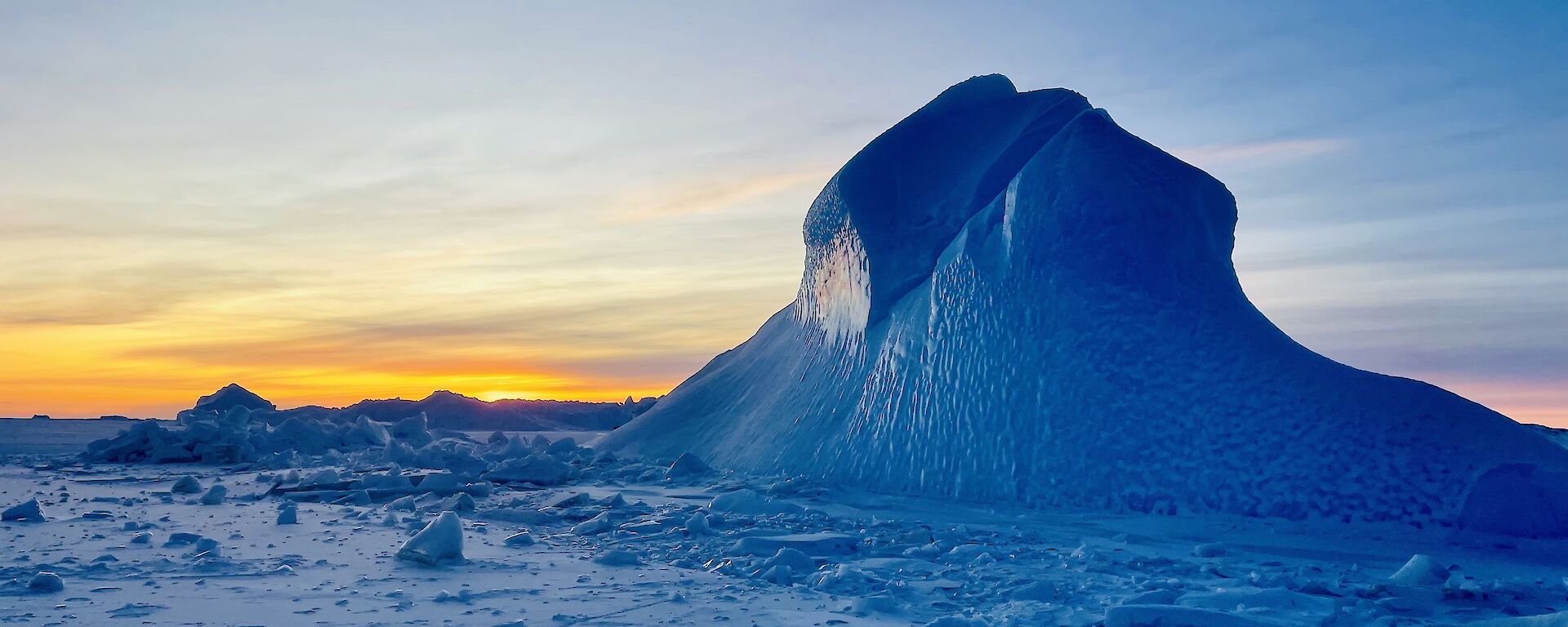 A tall blue iceberg pokes out of the frozen sea-ice. The distant horizon is shades of yellow and orange.