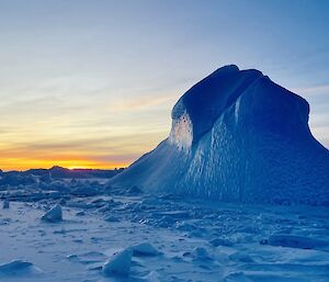 A tall blue iceberg pokes out of the frozen sea-ice. The distant horizon is shades of yellow and orange.